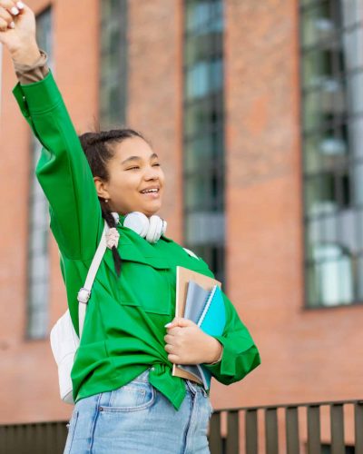 Happy mixed race female student celebrating success outdoors, successfully passed exam, standing at campus, raising hands and exclaiming with excitement, copy space