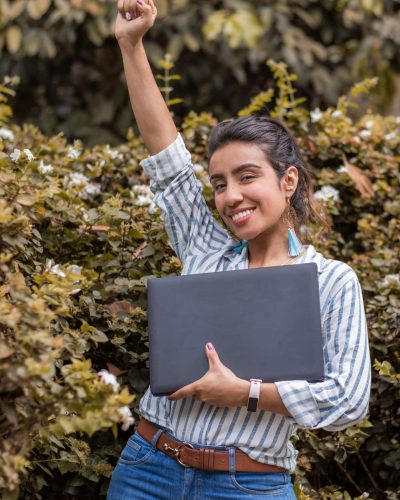 Exultant young woman celebrating, smiling and punching the air with her fists while holding her laptop. Successful woman concept